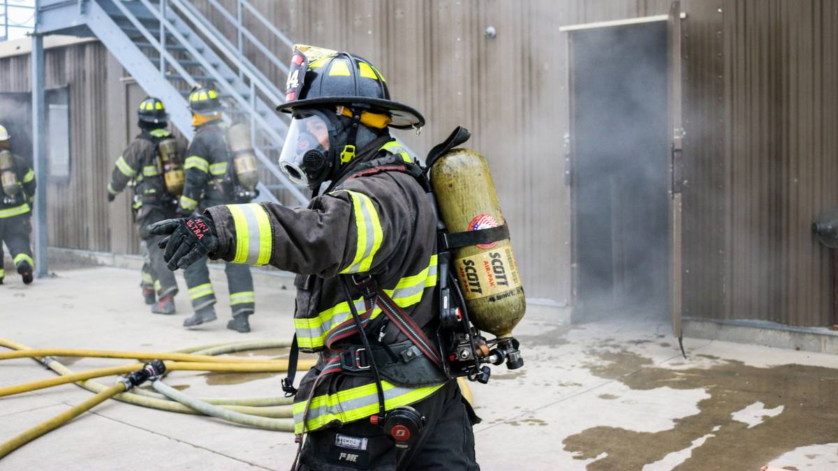 3 firefights walking off in distance next to burning building while one points in the foreground.