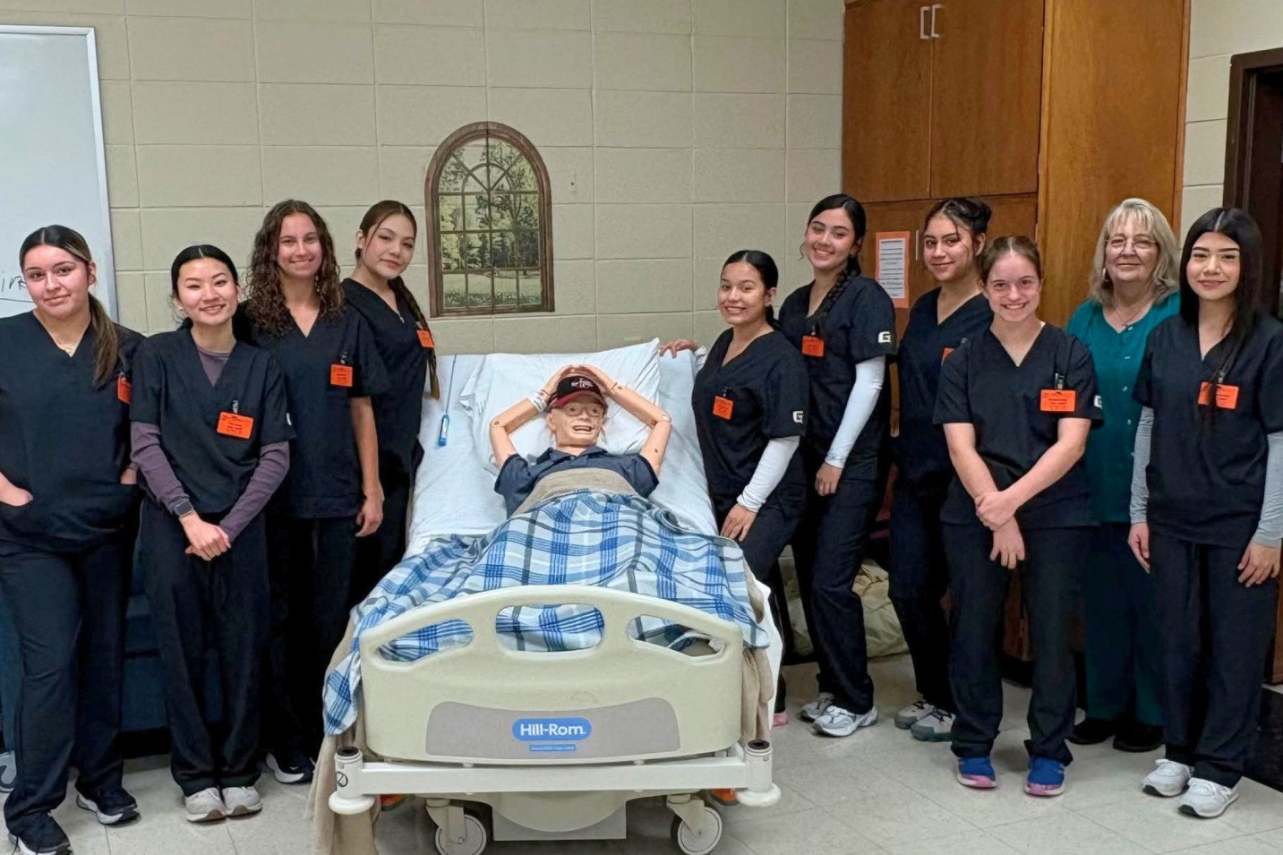 Group of healthcare students in navy scrubs standing around a patient simulation mannequin in a hospital bed during a clinical training session.
