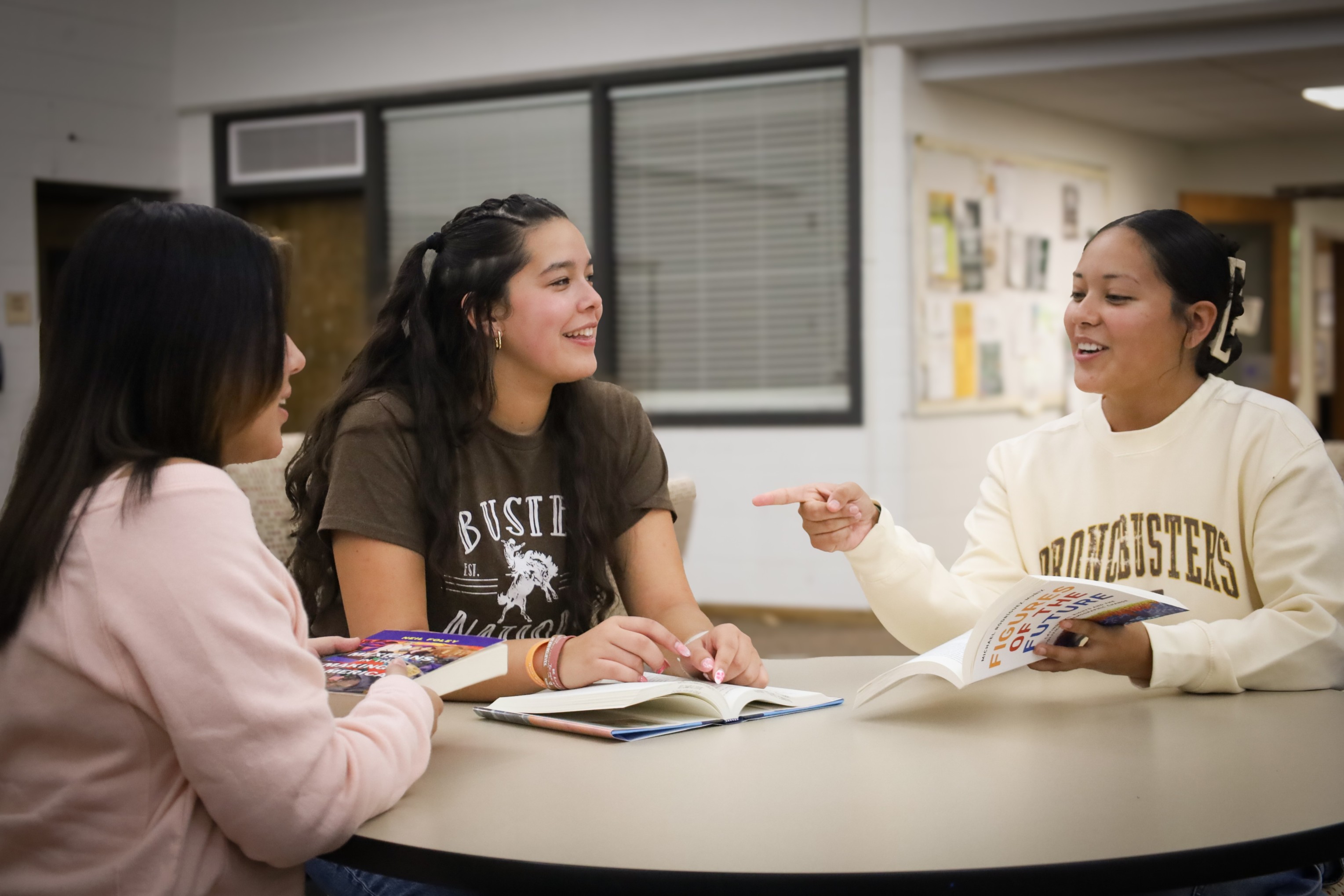Three people sitting at a table in a library, reading books and talking together.