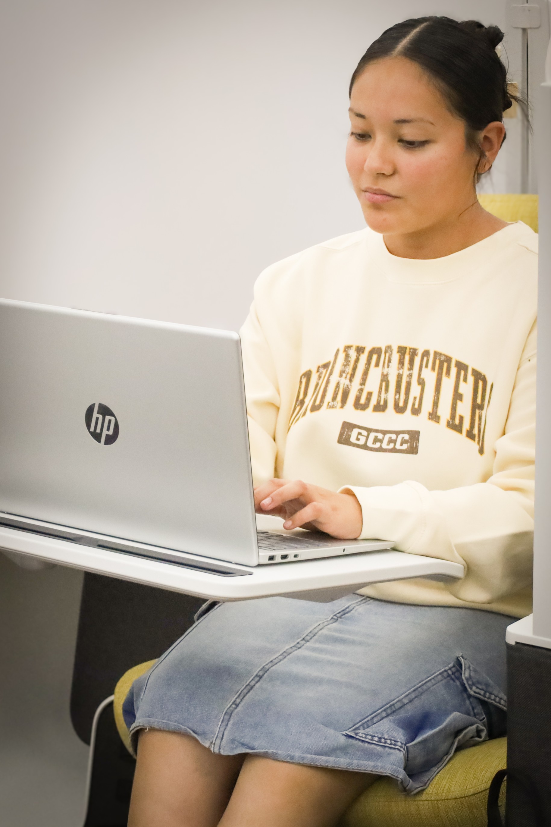 Person seated at a small desk using an HP laptop, wearing a “Broncbusters GCCC” sweatshirt.