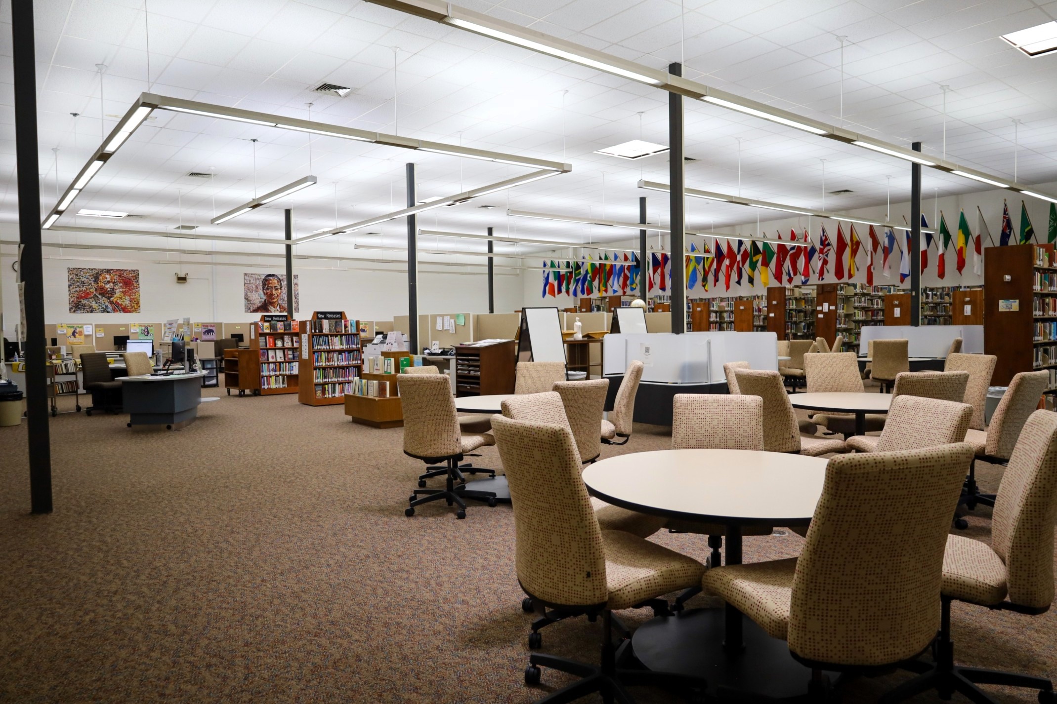 Modern library interior with round tables and rolling chairs, bookshelves, computer stations, and international flags hanging along the walls.