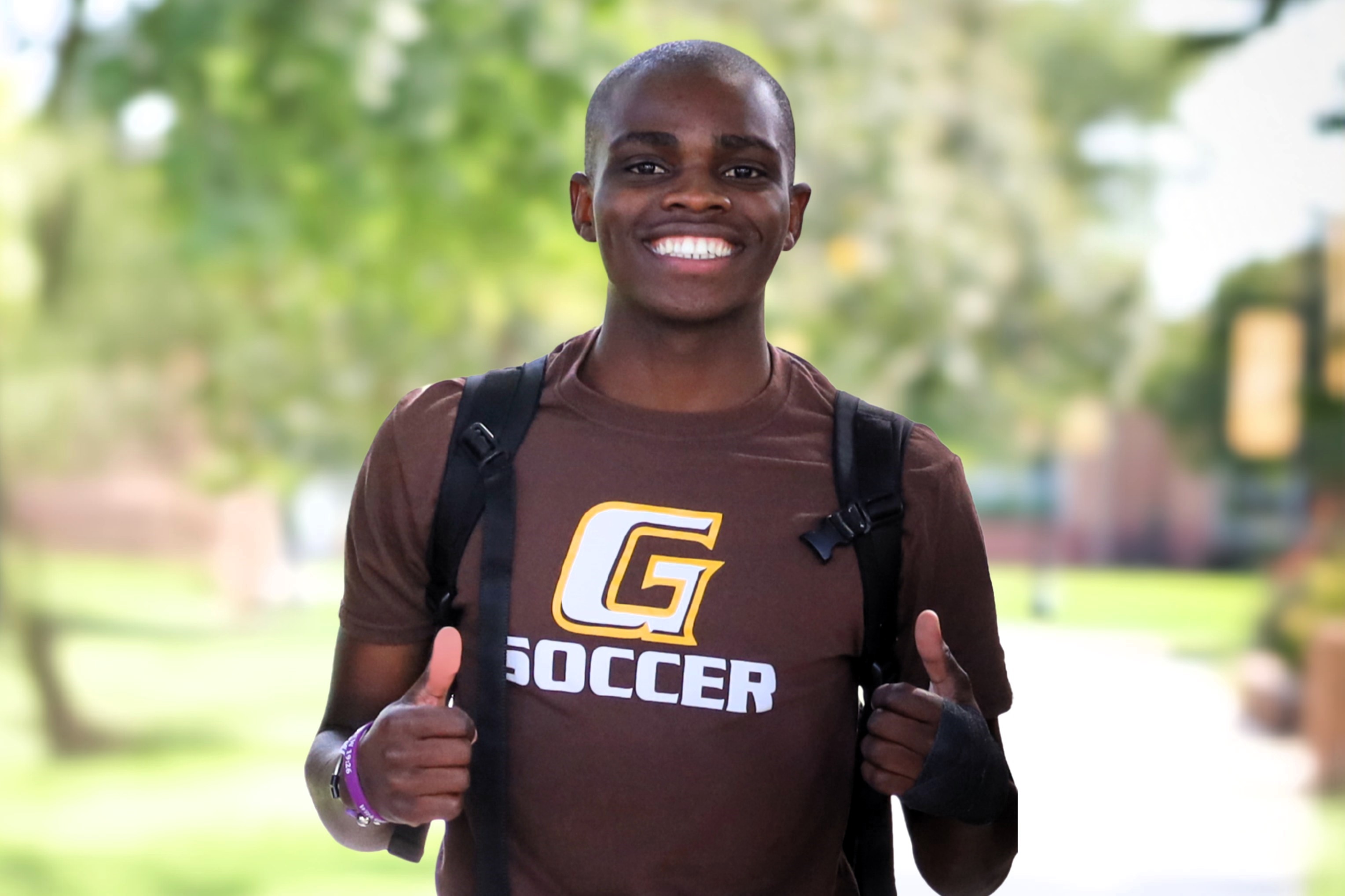 Student with backpack wearing a “G Soccer” T‑shirt standing outdoors on a campus walkway.