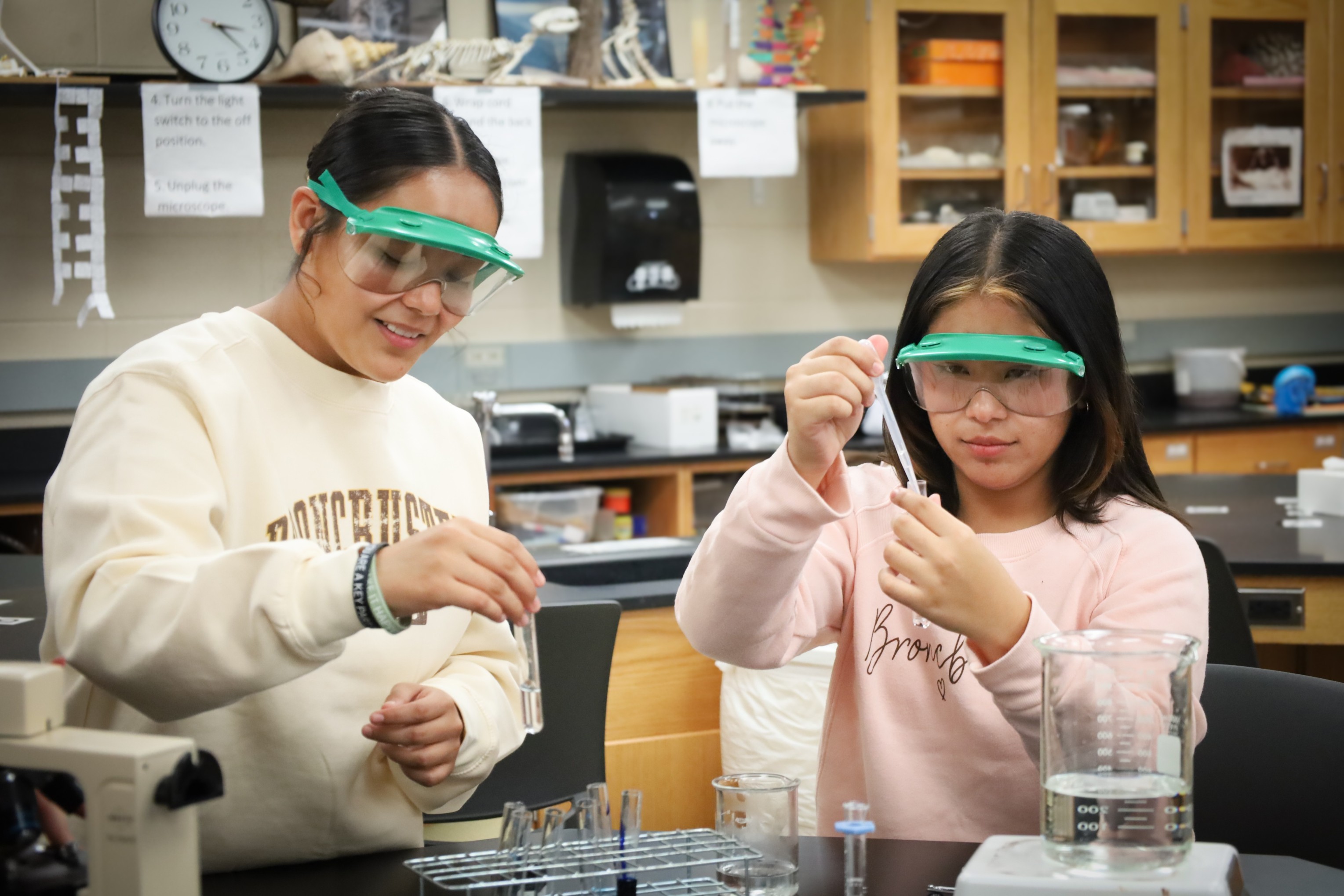 Two students wearing safety goggles conduct a science experiment in a classroom lab, using test tubes and beakers at a workbench.