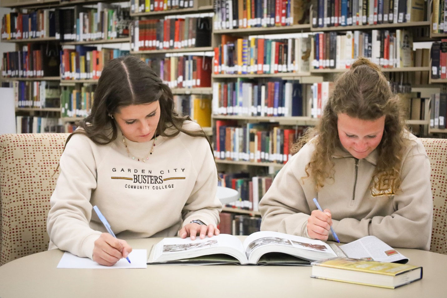 Two students studying in a library, writing in notebooks with open books on a table in front of them.