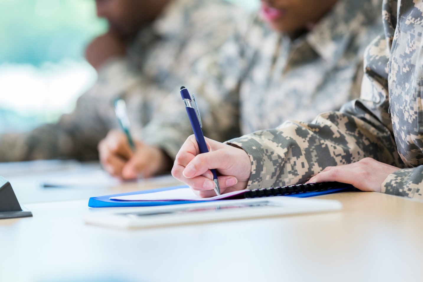 Close-up of people in camouflage uniforms writing on notebooks at a desk.