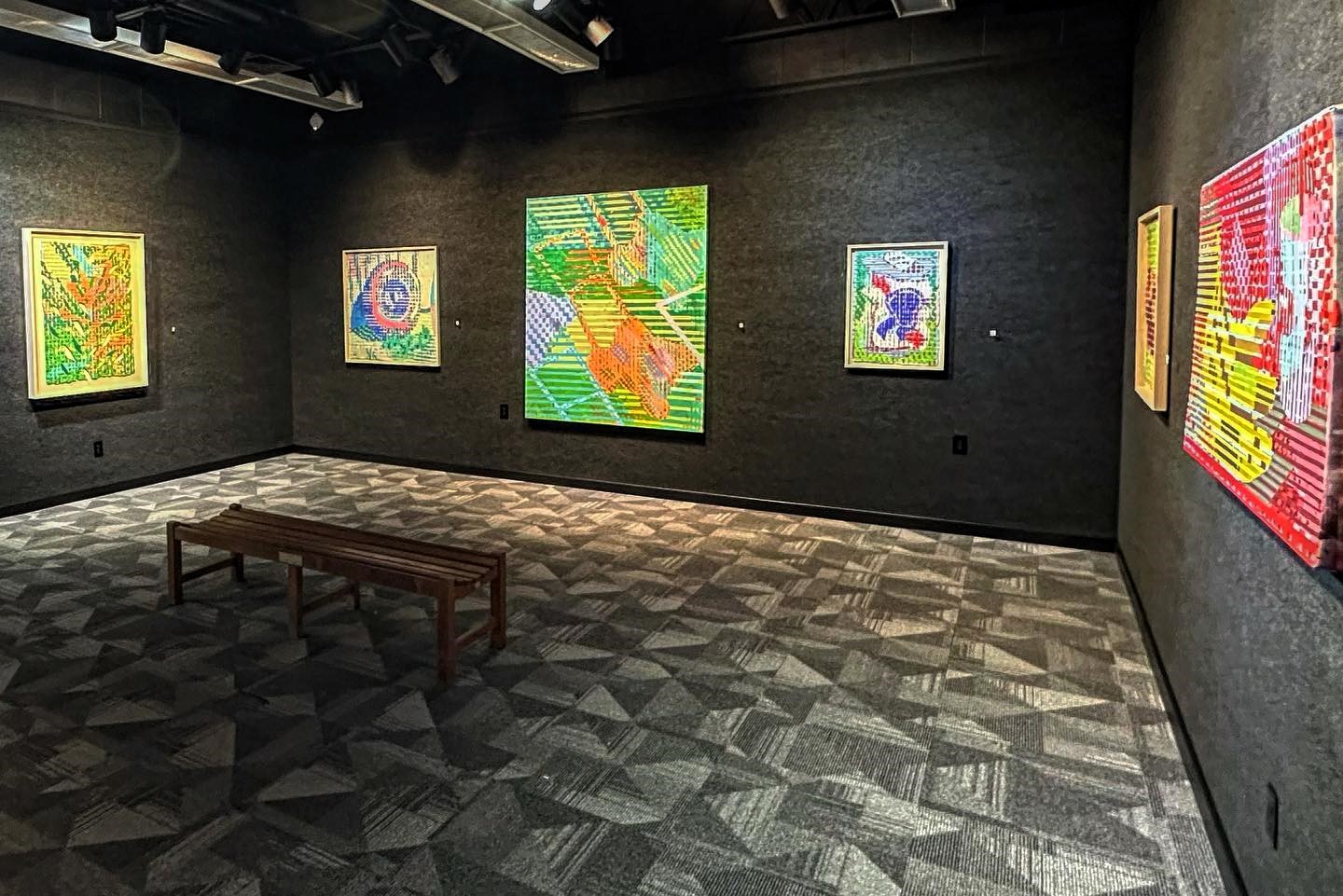 Art gallery interior with colorful framed artworks displayed on dark walls and a bench in the center.