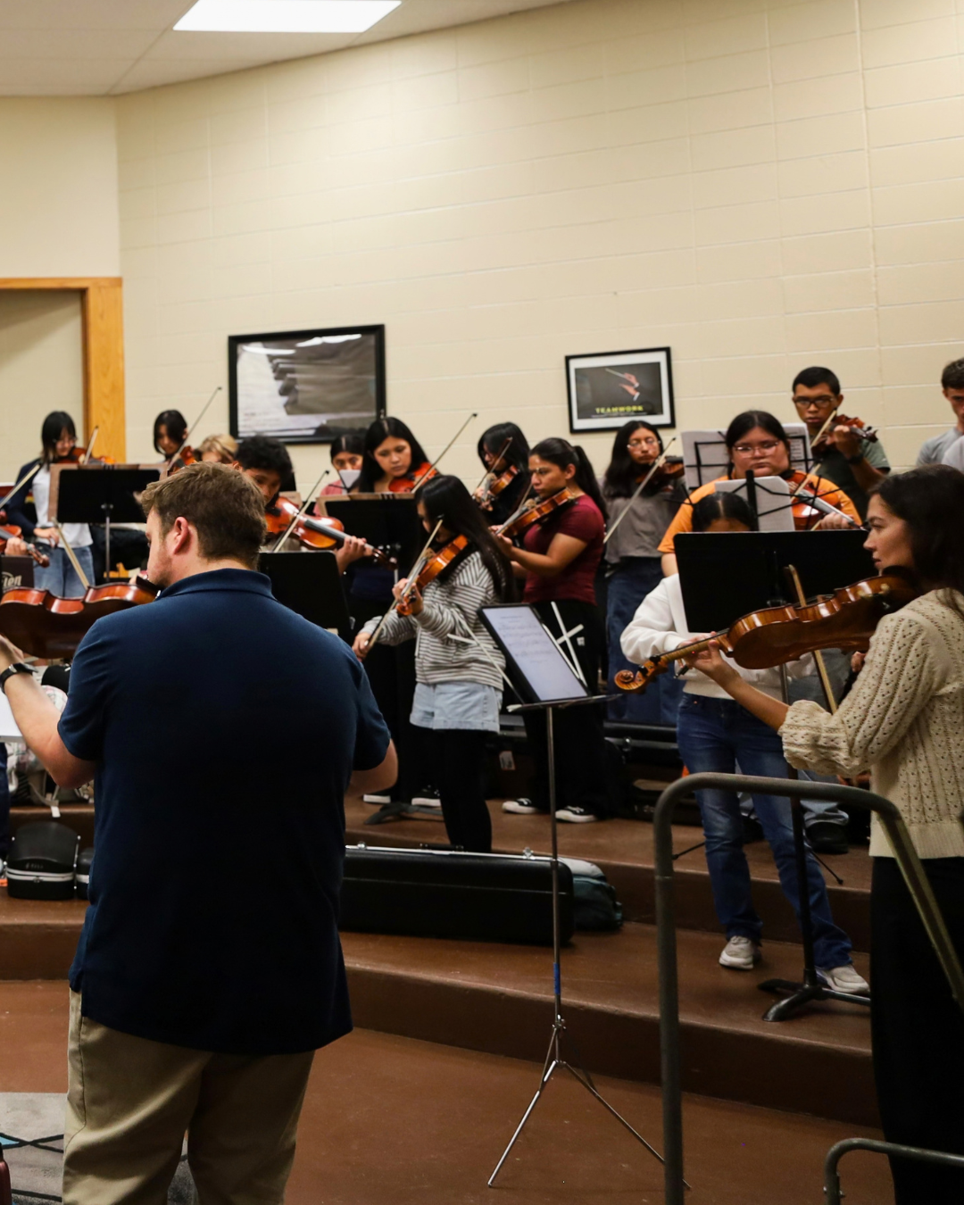 A group of young musicians rehearsing with violins under the guidance of a conductor in a classroom setting.