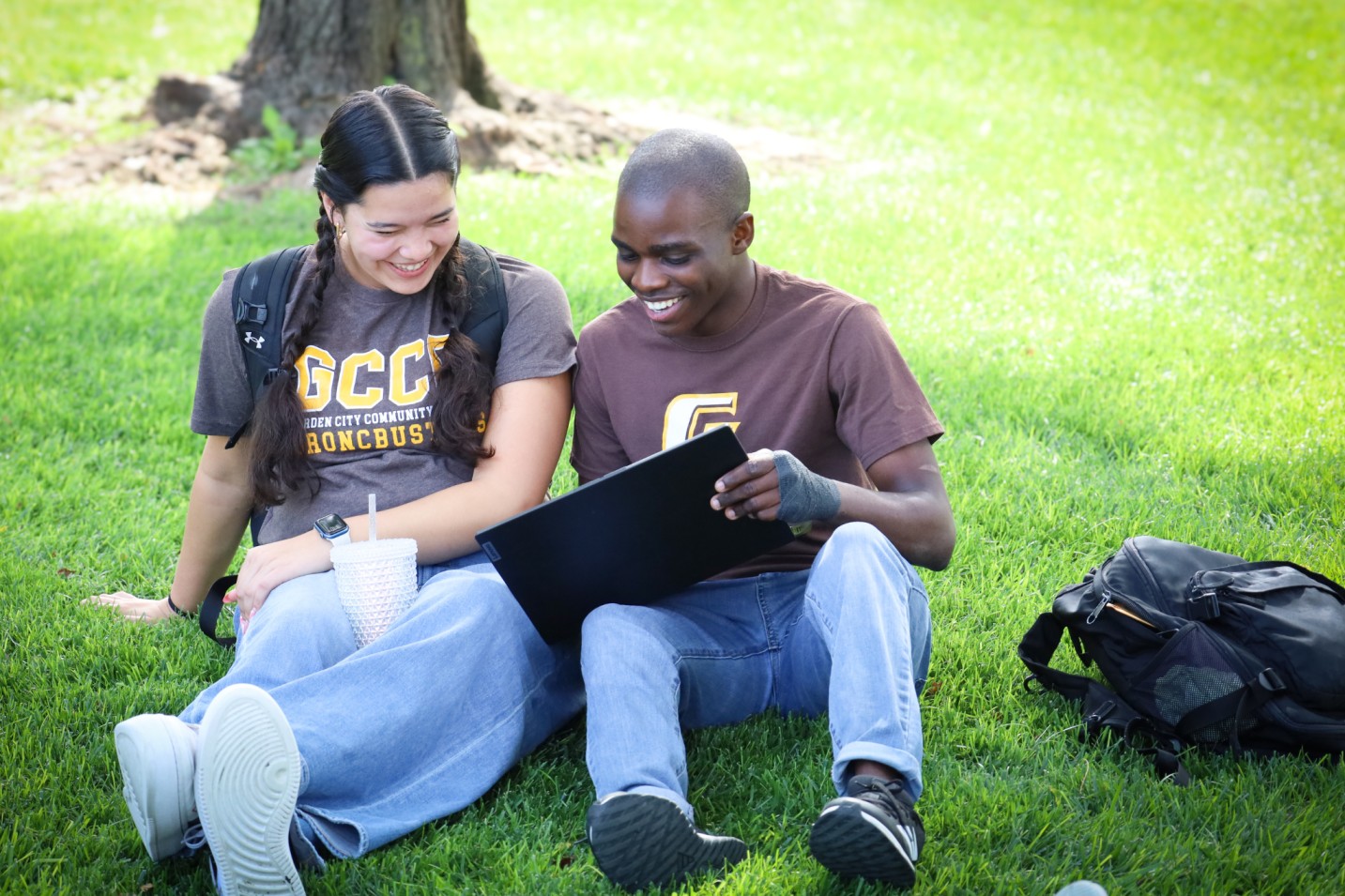 Students sitting on grass outside, looking at a laptop together with backpacks nearby.