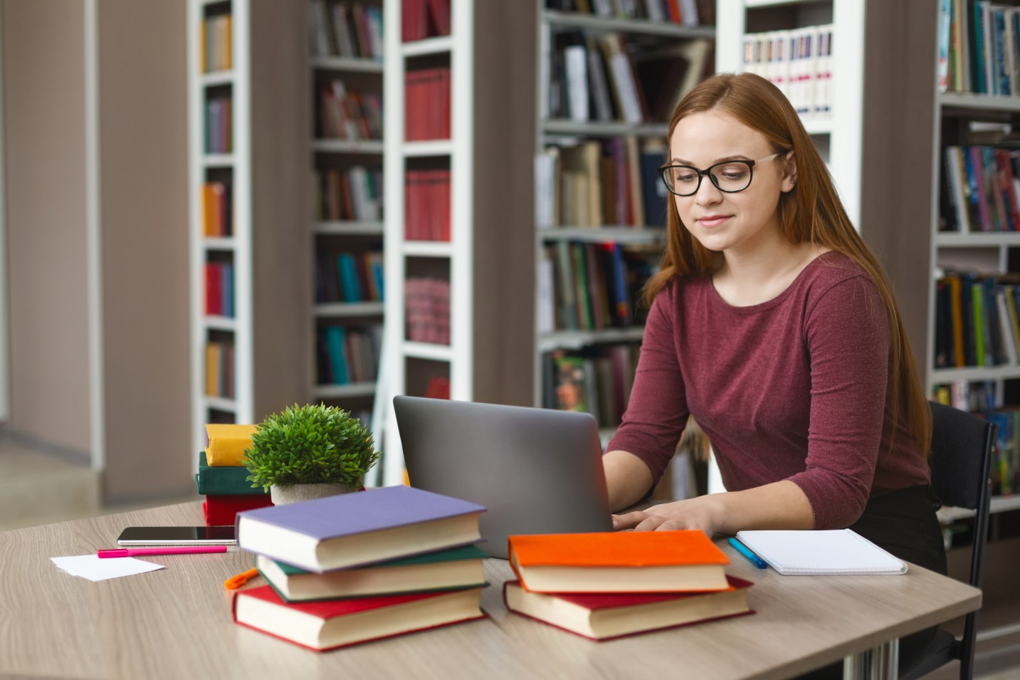 Person working on a laptop at a table with stacked books in a library.
