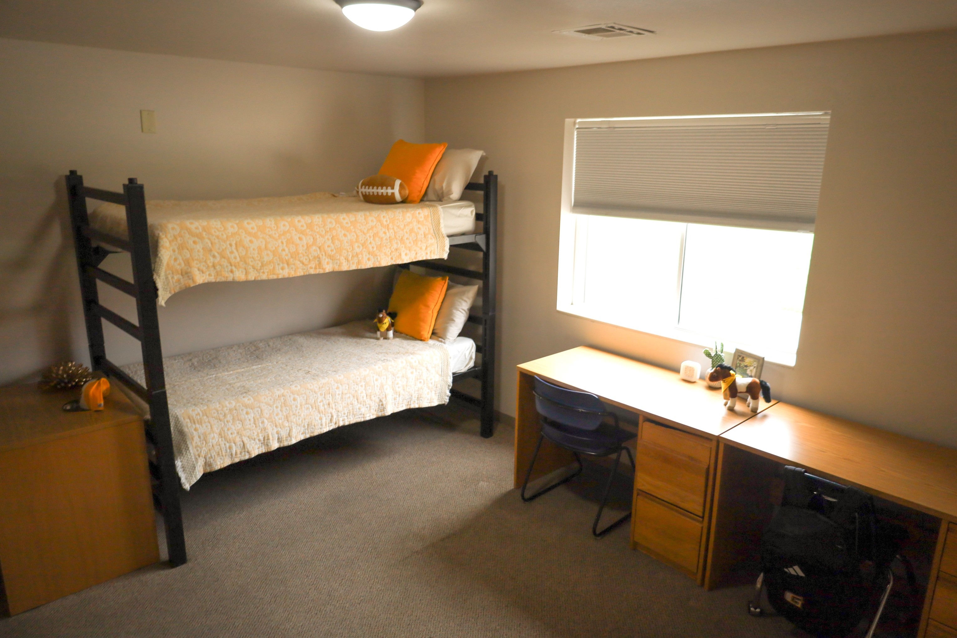 Bedroom with bunk bed, wooden desk and chair by a window with blinds, and small decorative items on the desk.