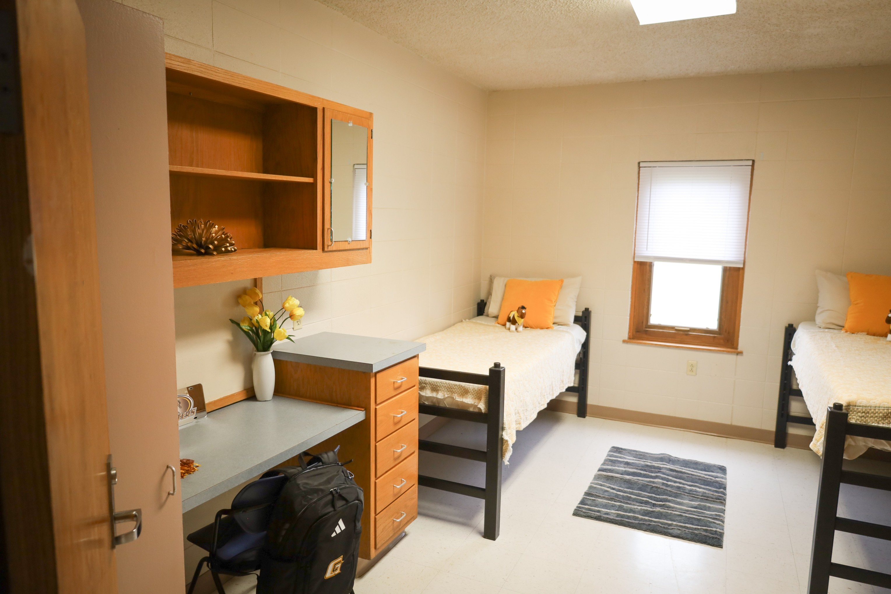 Dorm-style bedroom with two single beds, wooden desk and shelves, window with blinds, and small rug on tile floor.
