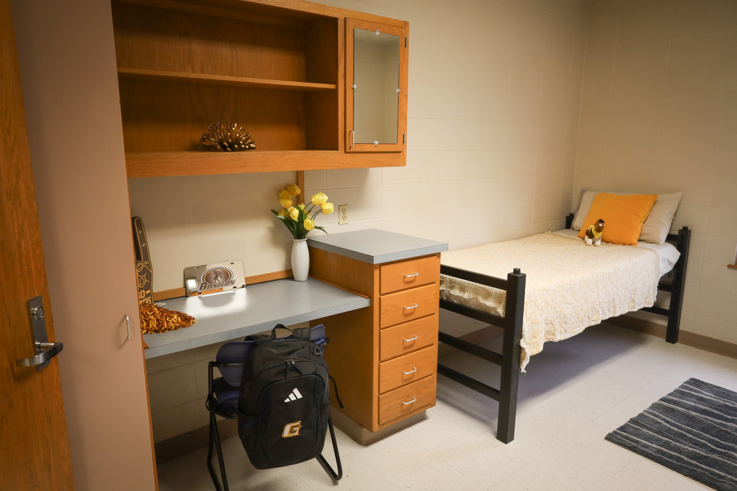 Dorm-style bedroom with single bed, wooden desk and shelving unit, backpack under desk, and small rug on tile floor.