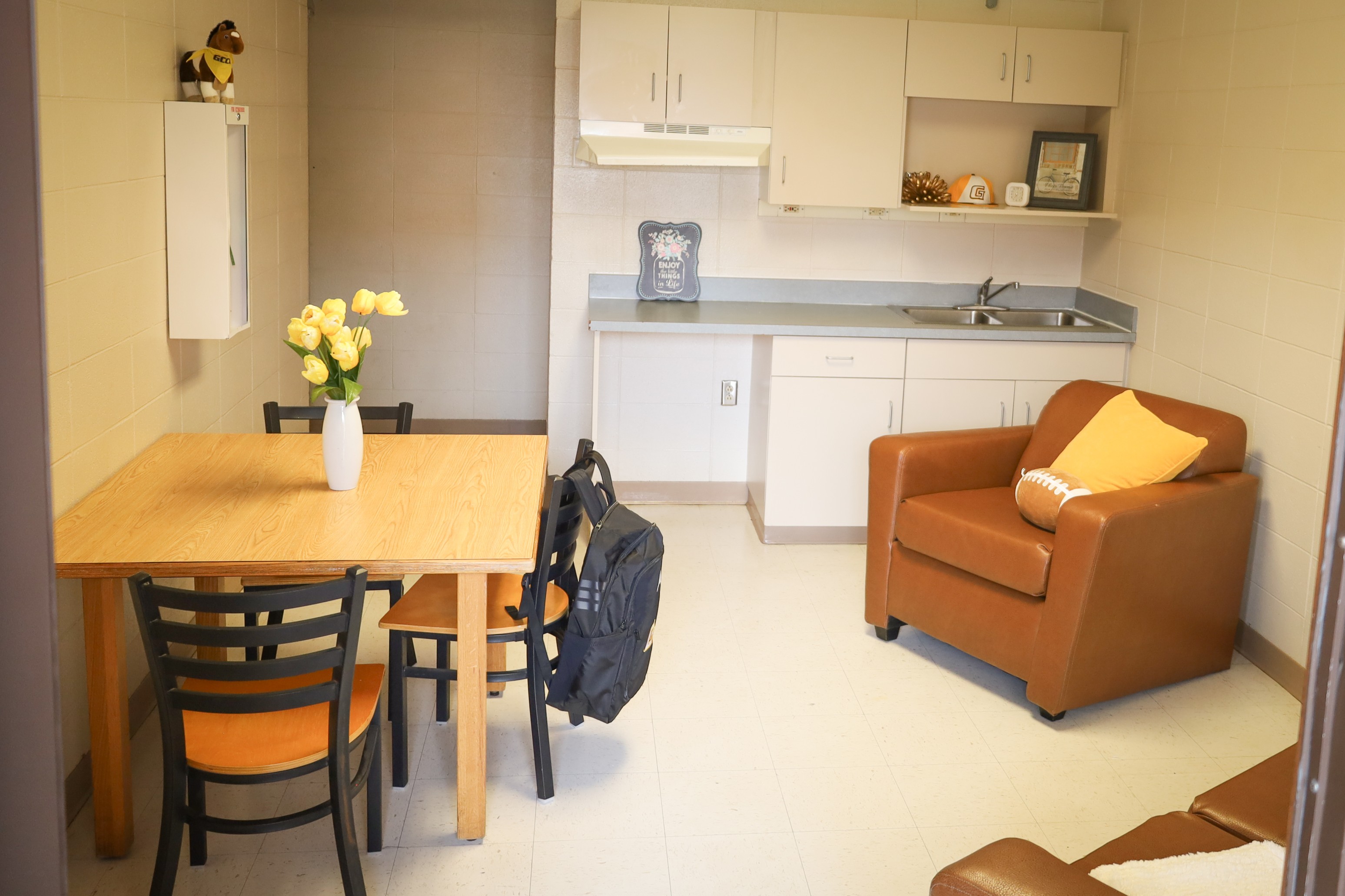 Small kitchenette and dining area with table and chairs, sink and cabinets, and a cushioned chair with accent pillow.