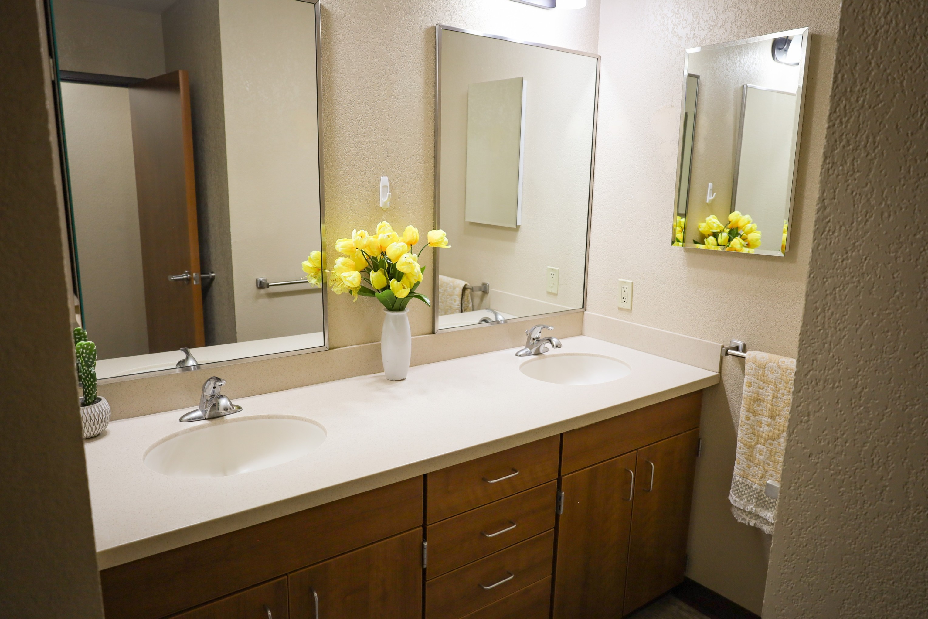 Bathroom vanity with double sinks, large mirrors, wood cabinets, and a vase of yellow flowers on the counter. Provide your feedback on BizChat