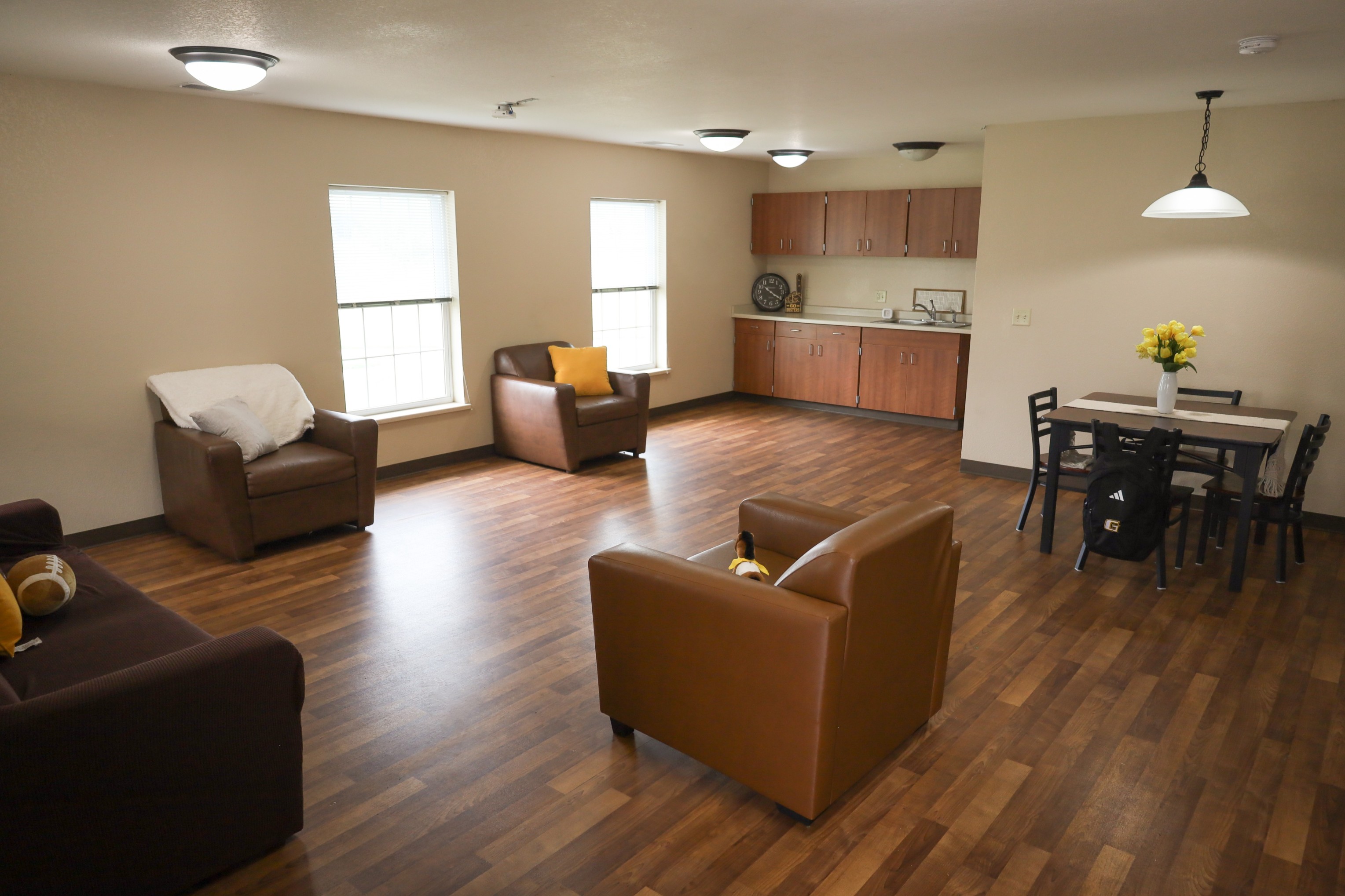 Spacious living area with brown chairs, wood flooring, kitchenette along the wall, and a small dining table with chairs.