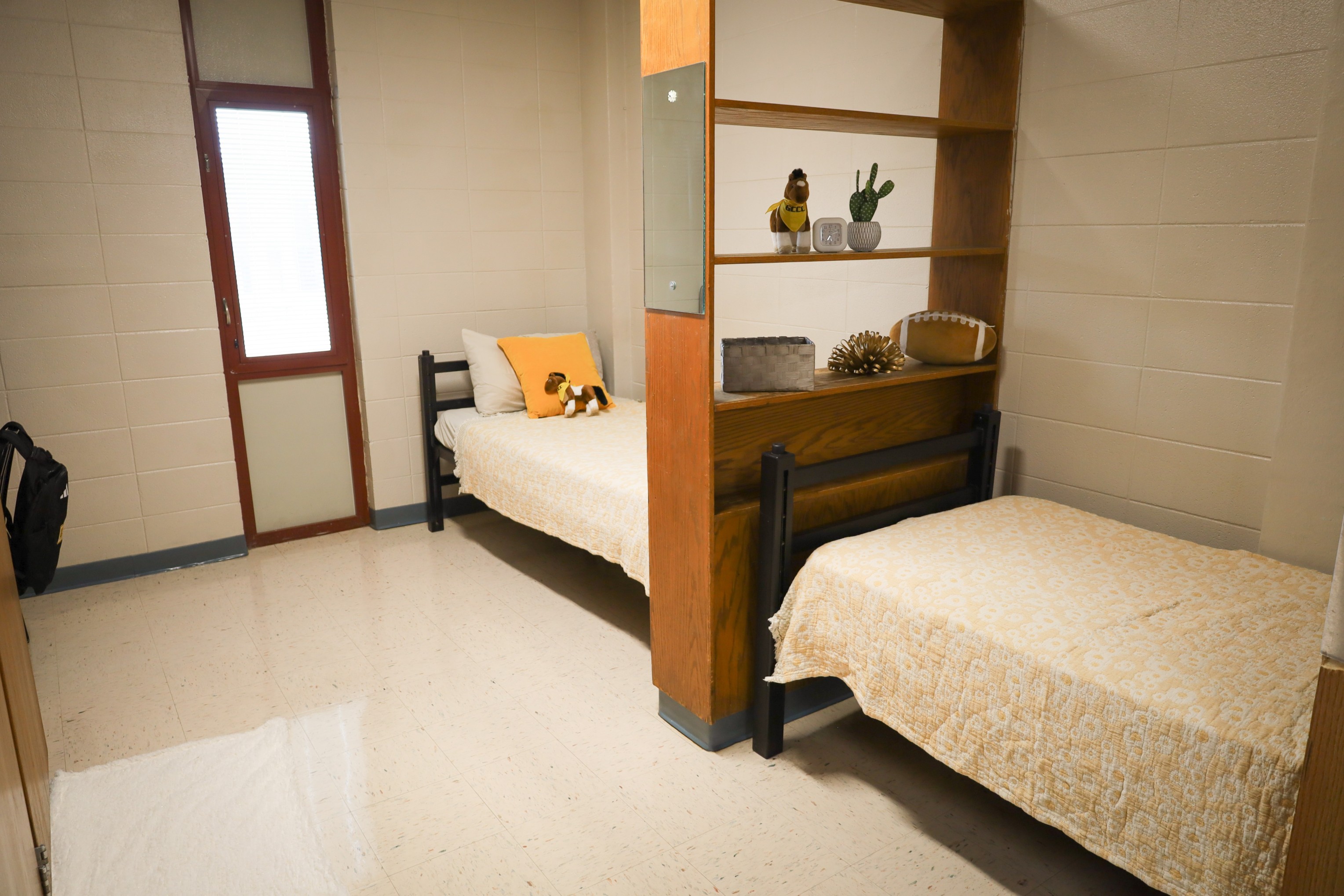 Dorm-style room with two single beds separated by a wooden shelving divider, window with blinds, and light tile floor.