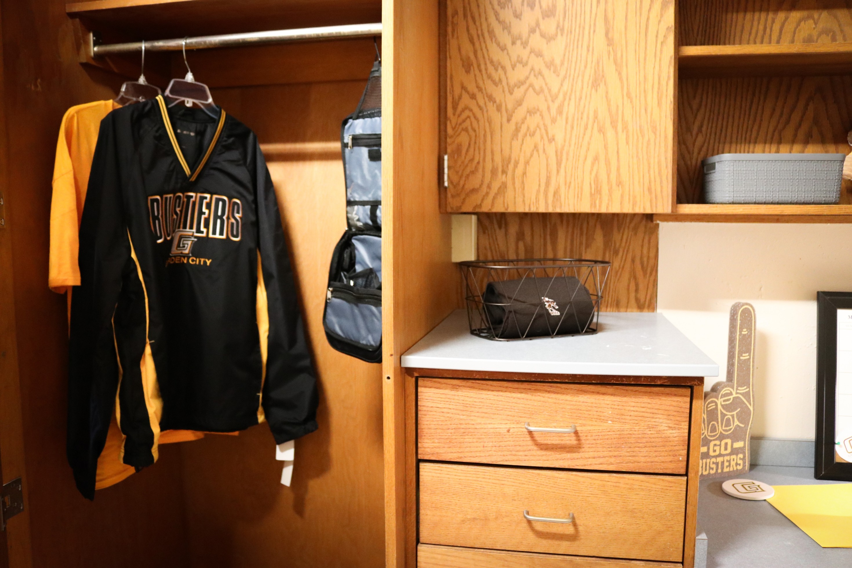 Open dorm closet with hanging clothes and storage shelves next to a wooden dresser and desk area.
