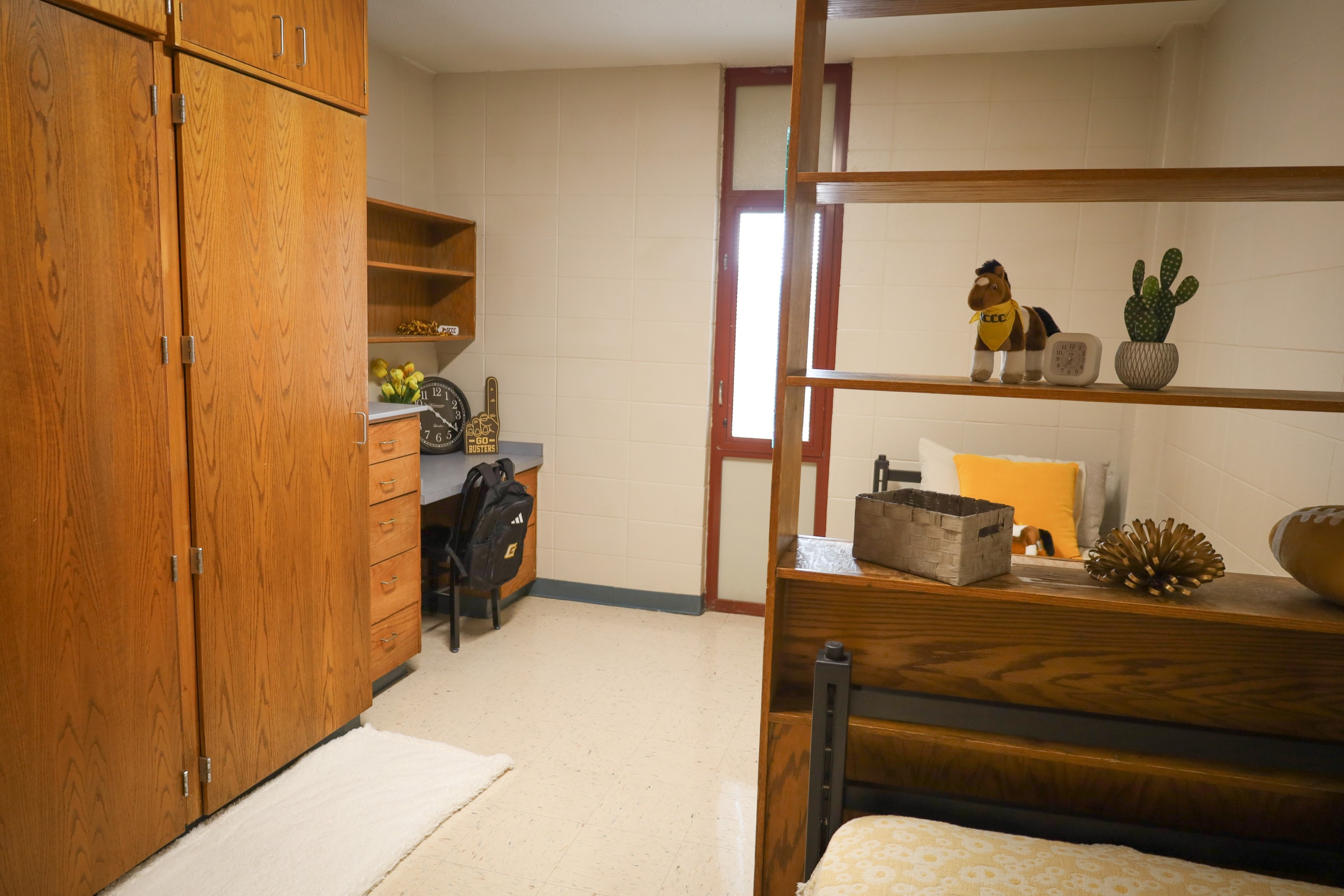 Dorm-style room with two beds separated by a wooden shelving divider, desk area, wardrobe, and window with blinds.