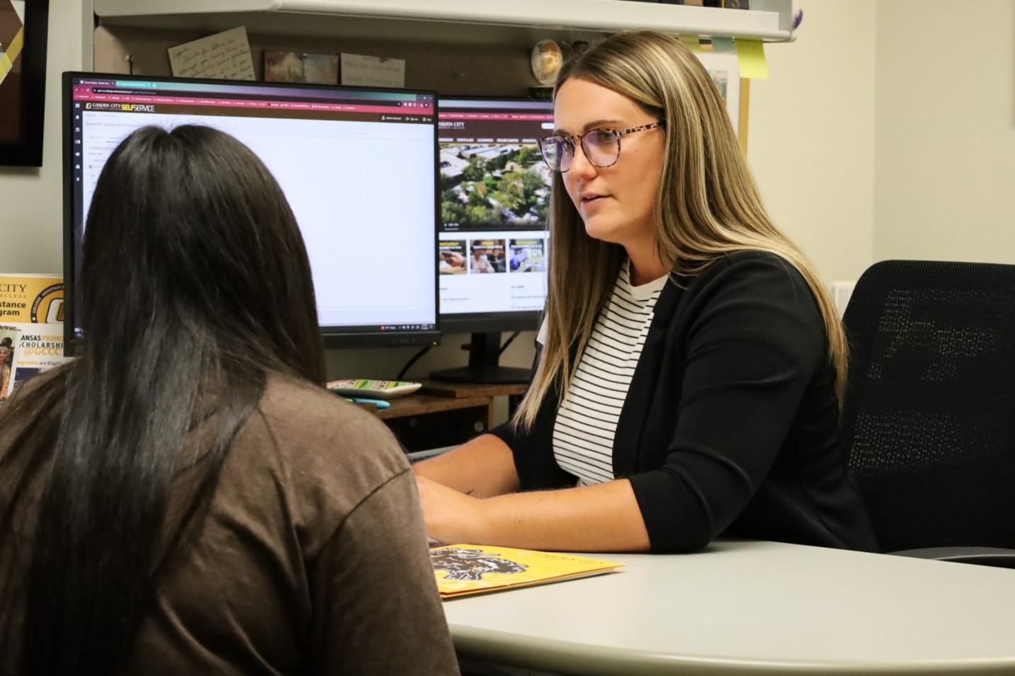 Student and staff member meeting at a desk in an office, reviewing information on dual computer monitors.