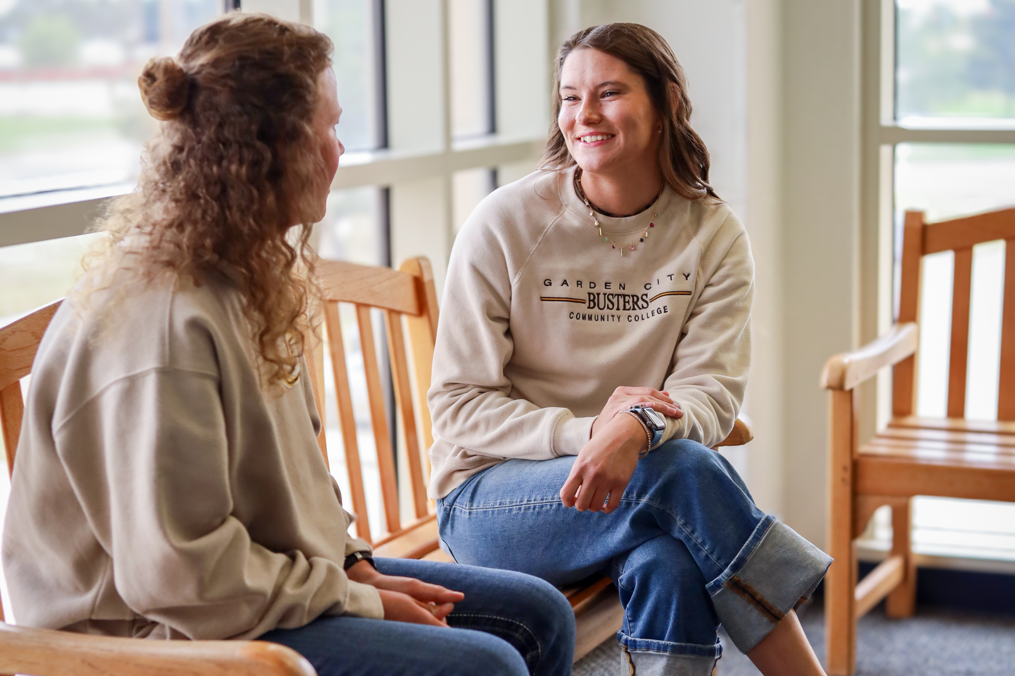 Two students seated on benches in a campus common area, talking near large windows.