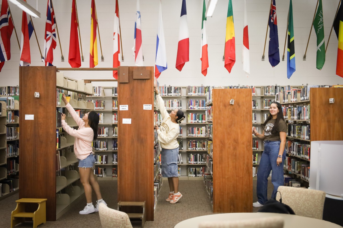 Copilot said: Students browsing and selecting books from library shelves beneath rows of international flags.