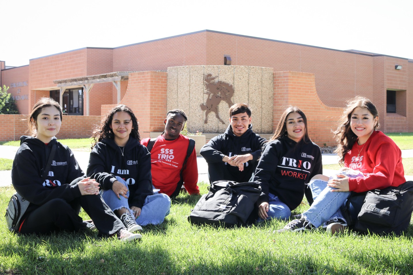 Group of students wearing TRIO shirts sitting on grass in front of a school building.