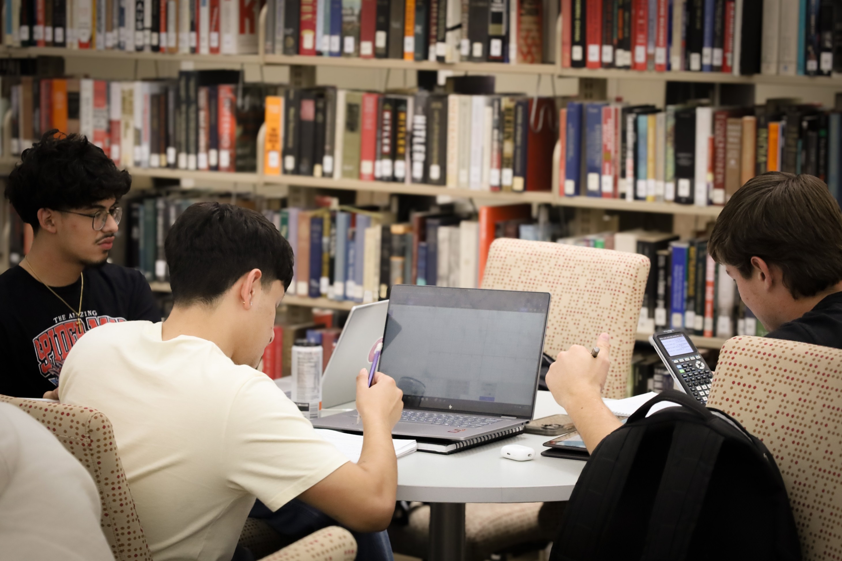 Students studying together at a library table with laptops, notebooks, and a calculator.